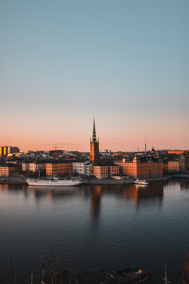 Vista de la nueva guía de viaje de Estocolmo al atardecer, con la iglesia de Riddarholmen iluminada por la luz dorada y reflejos en el agua, mostrando la arquitectura histórica junto al muelle.