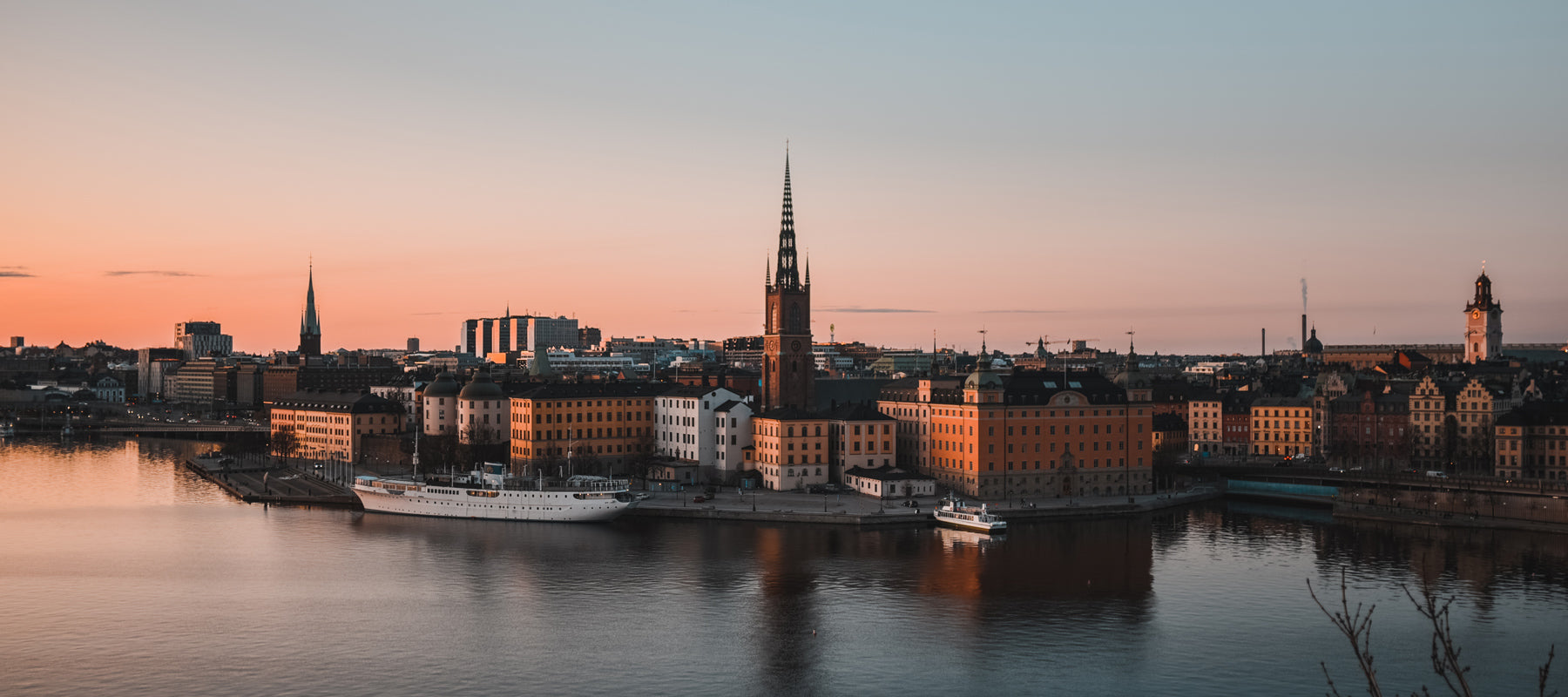 Vista de la nueva guía de viaje de Estocolmo al atardecer, con la iglesia de Riddarholmen iluminada por la luz dorada y reflejos en el agua, mostrando la arquitectura histórica junto al muelle.