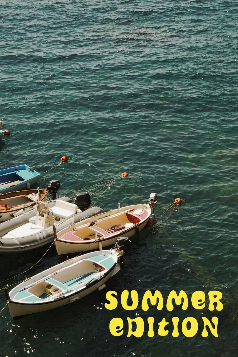 Vista de la costa italiana en verano, con aguas turquesas y pequeñas barcas de pescadores flotando cerca de un pintoresco pueblo costero. Obtén un 20% de descuento por la compra de una guía, descuento canjeable en el siguiente pedido.