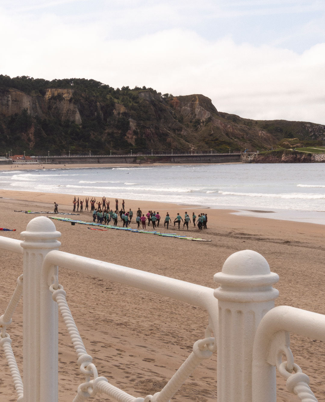 Camino costero por las salinas entre montaña y playa con vegetación frondosa y mar al fondo y surfistas entrenando.