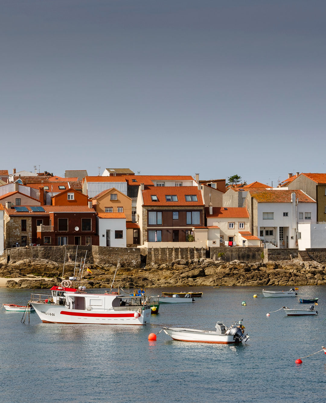 Vista de un puerto pesquero en Corrubedo con barcos amarrados y casas bajas de fondo.

